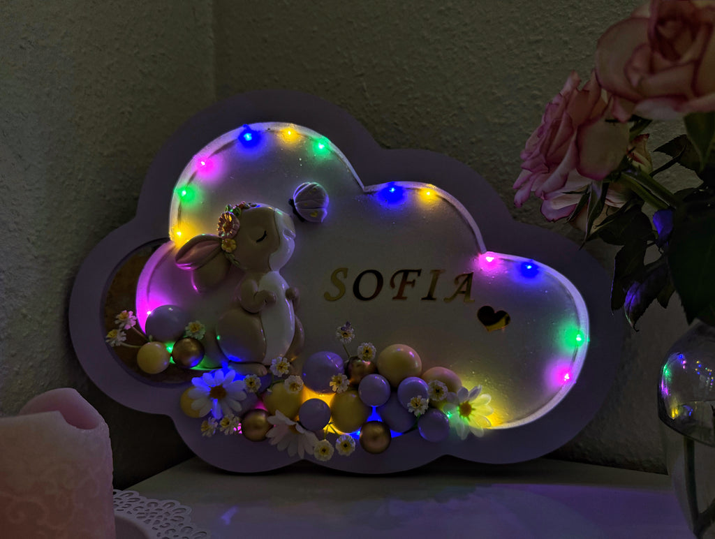 Decorative cloud-shaped plaque with LED lights, teddy bear, and 'SOFIA' text on a dark background.